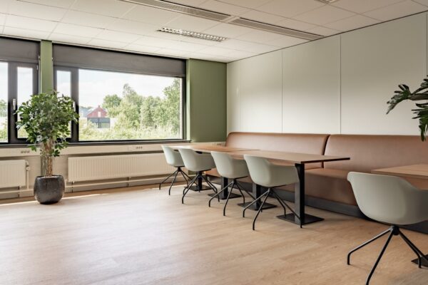 Moderne, lichte zithoek met houten tafels, grijze stoelen en een lange bruine bank in een ruimte aan de Burgemeester Verderlaan.